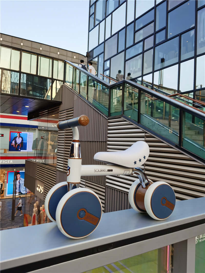 Retrospec Cricket Baby Walker Balance Bike displayed on urban ledge with modern architecture in background.