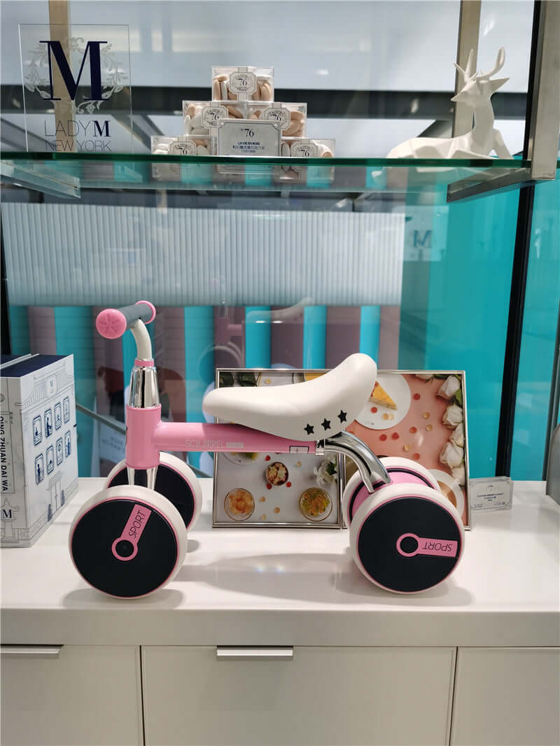 Pink Retrospec Cricket Baby Walker Balance Bike with four wheels displayed on a shelf.